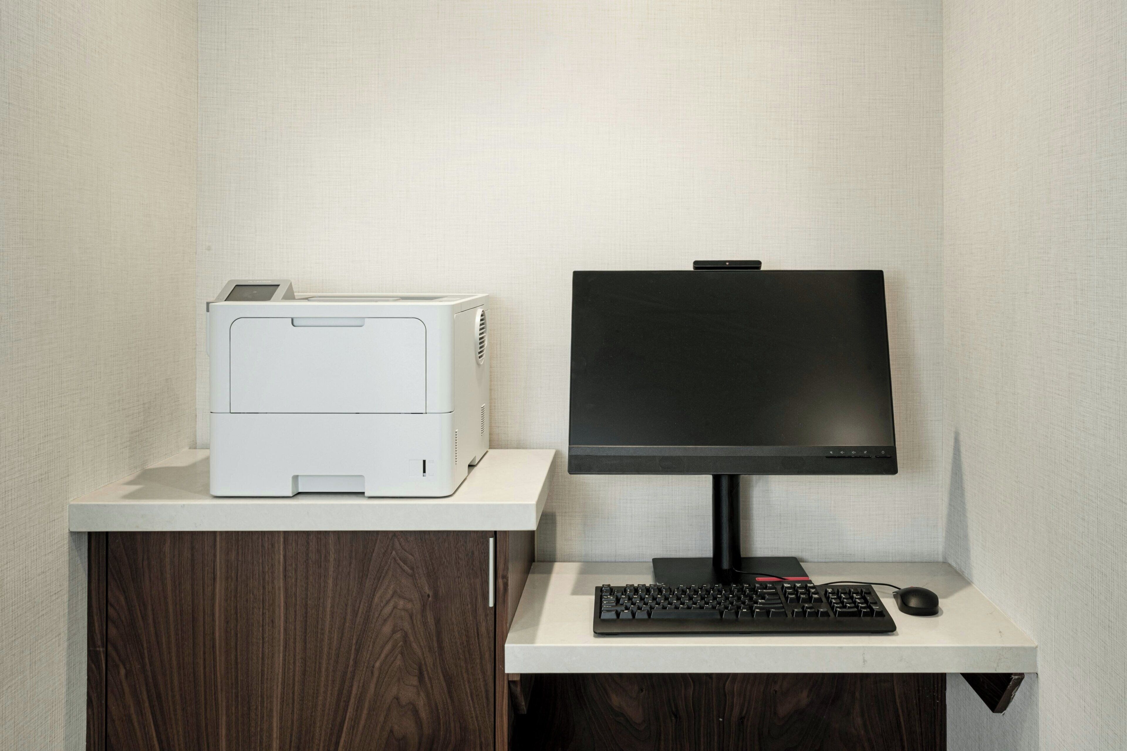 Organização de escritório compacta, com impressora, monitor, teclado e rato sobre uma secretária branca, com uma parede texturizada ao fundo.