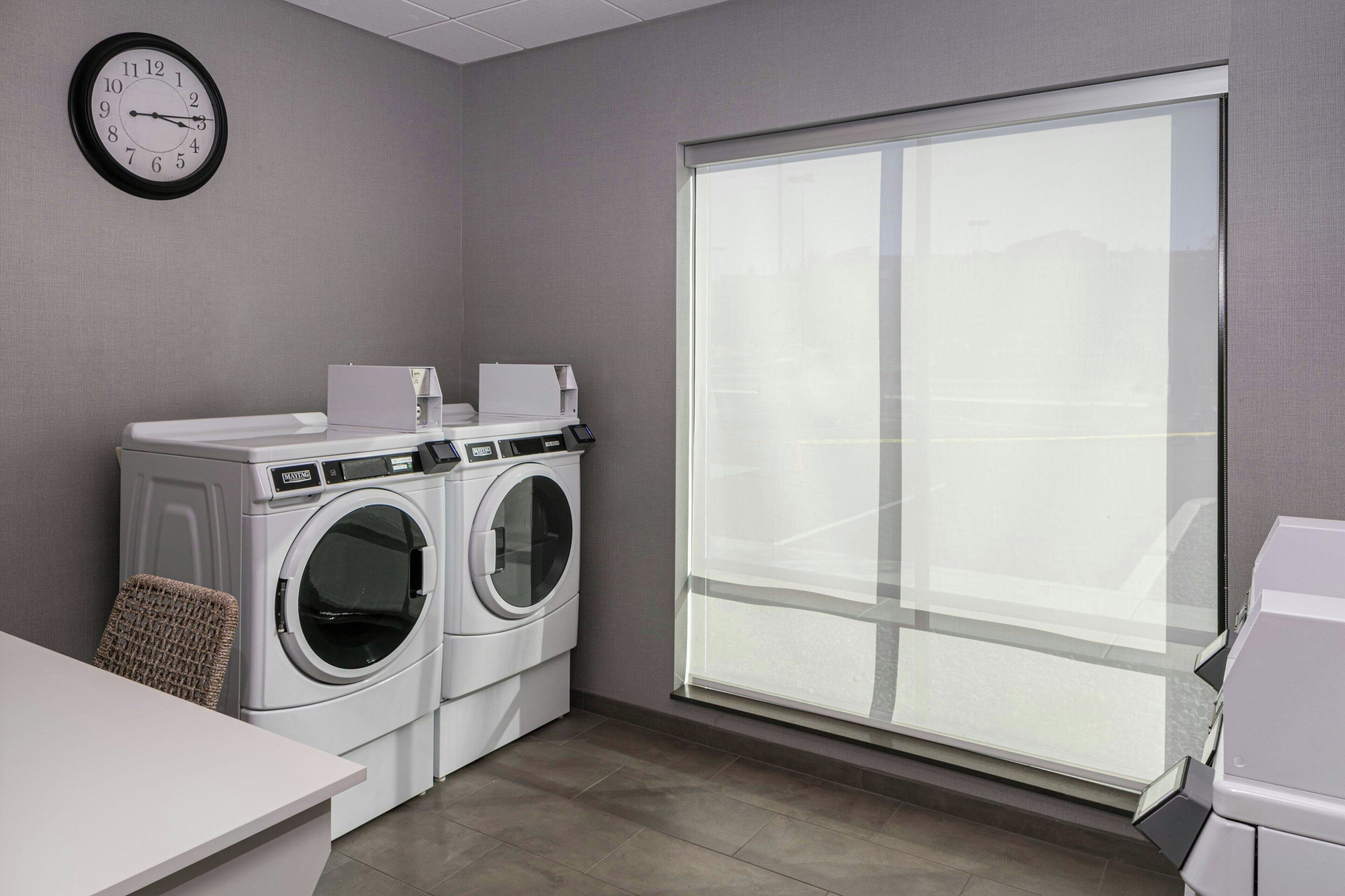 A laundry room with two front-loading washing machines, a wall clock, and a large window.