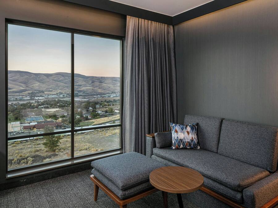 A modern hotel room with a large window offering a scenic view of hills and a town. There is a gray sofa with a patterned pillow and a small wooden table.