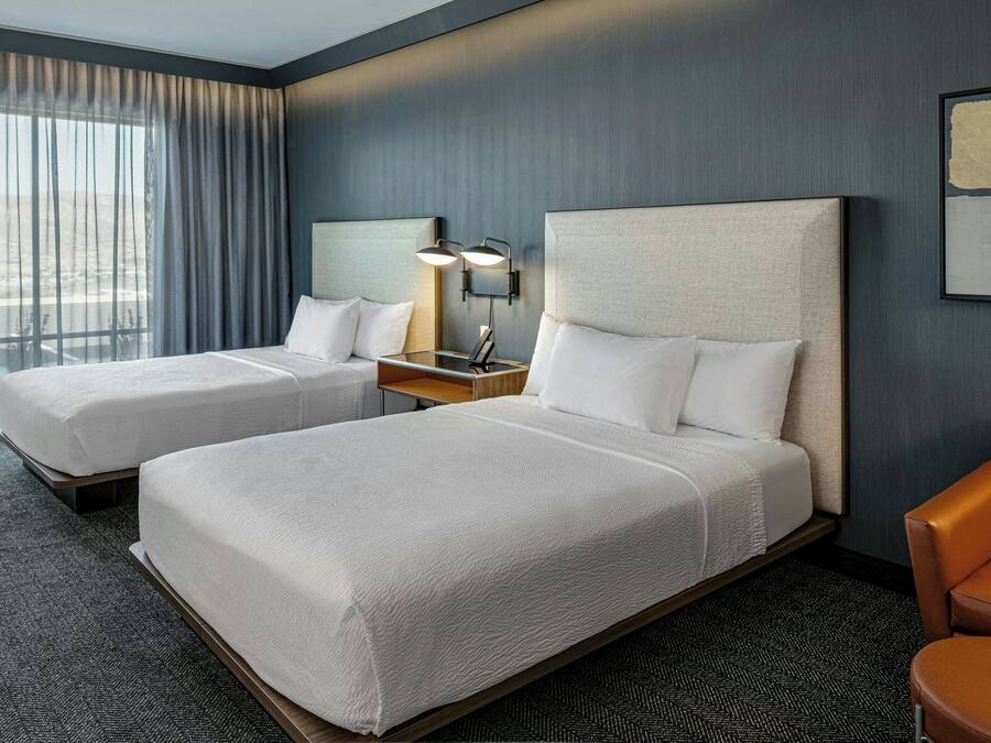 A hotel room with two beds, a nightstand, a lamp, and a view of the city.