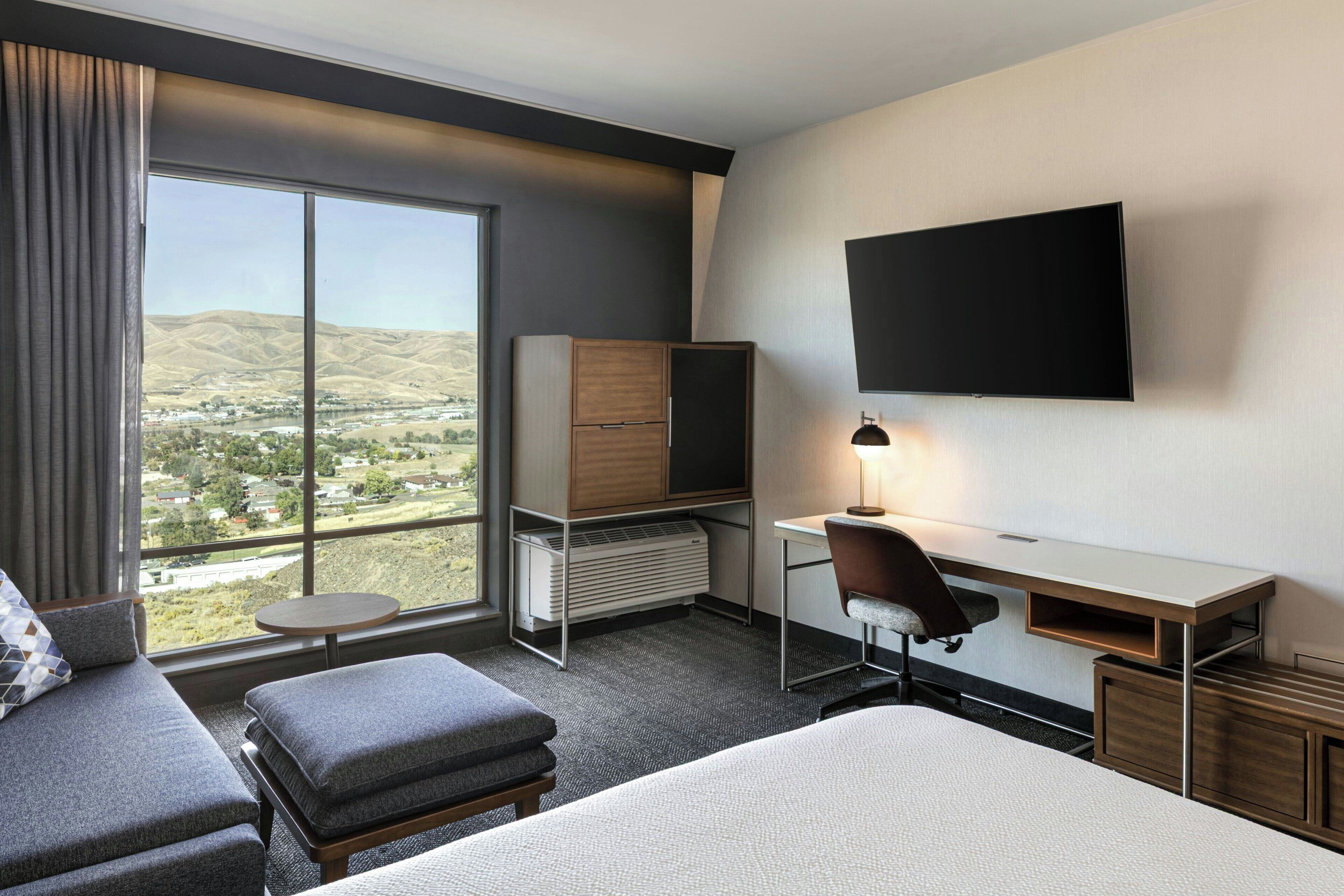 A hotel room with a large bed, a desk with a chair, a television mounted on the wall, and a view of a valley through a large window.