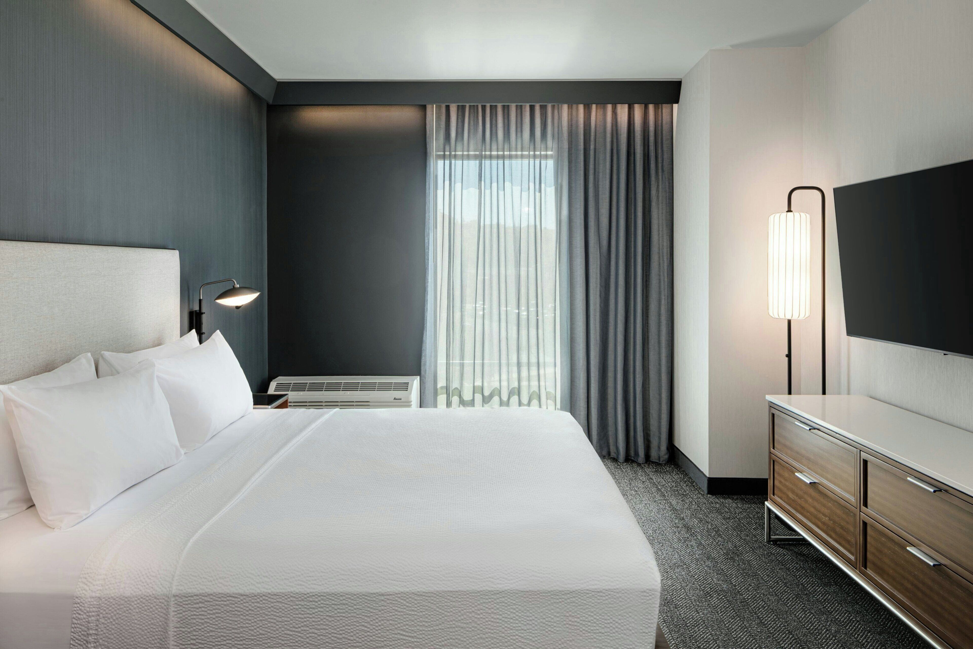 Une chambre d’hôtel avec un grand lit, une commode, un téléviseur à écran plat et une fenêtre avec des rideaux.