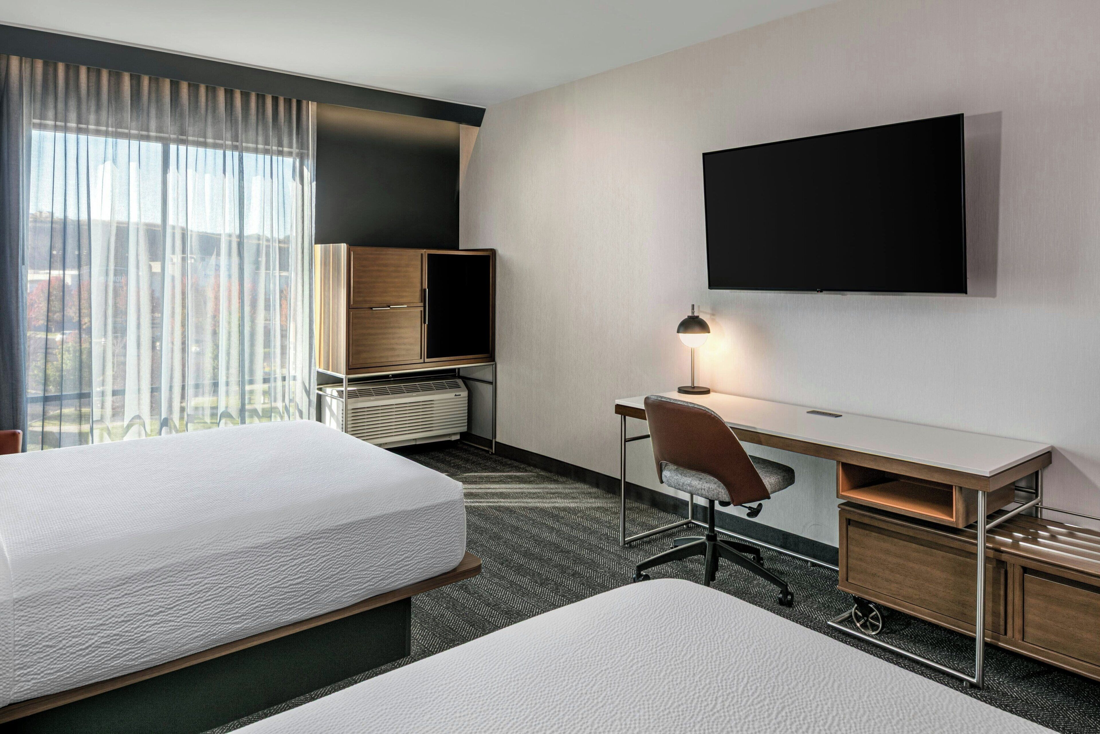 A hotel room with two beds, a desk with a chair, a flat-screen TV mounted on the wall, and a window with sheer curtains.