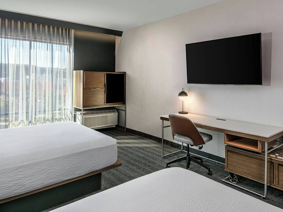 A hotel room with two beds, a desk with a chair, a flat-screen TV mounted on the wall, and a window with sheer curtains.