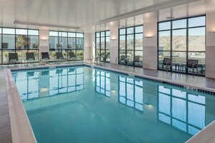 An indoor swimming pool with clear glass walls, lounge chairs, and a view of the mountains.