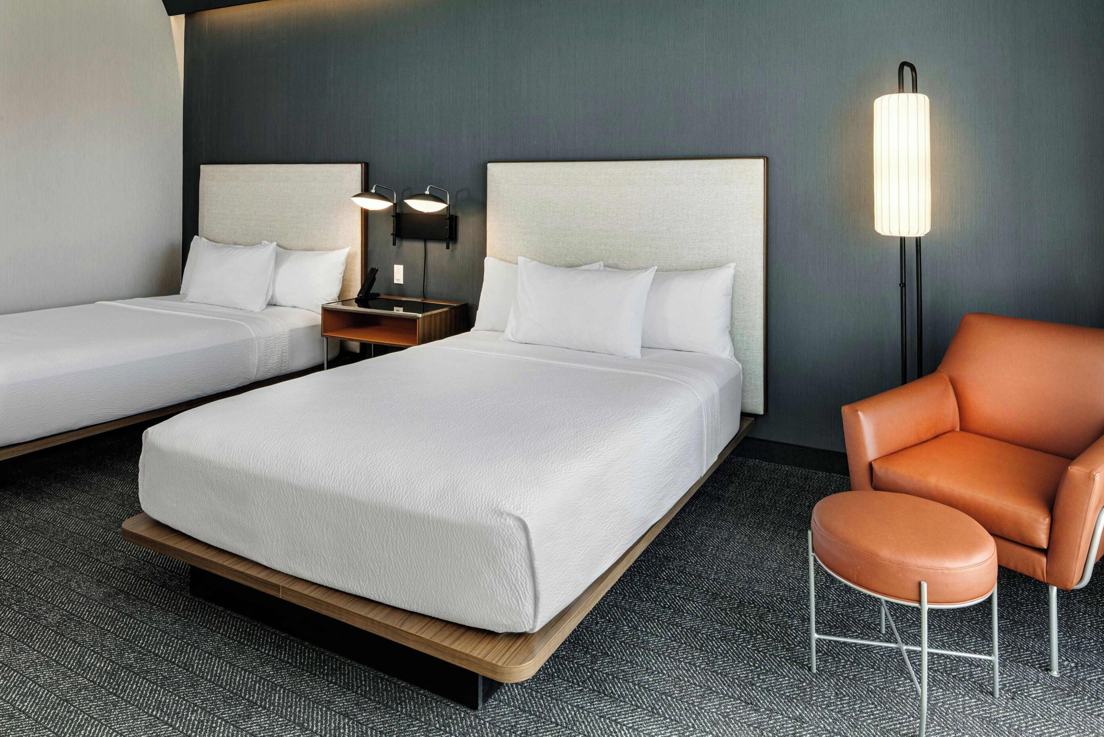 Hotellihuone, jossa on kaksi sänkyä, yöpöytä, lamppu ja nahkainen nojatuoli.