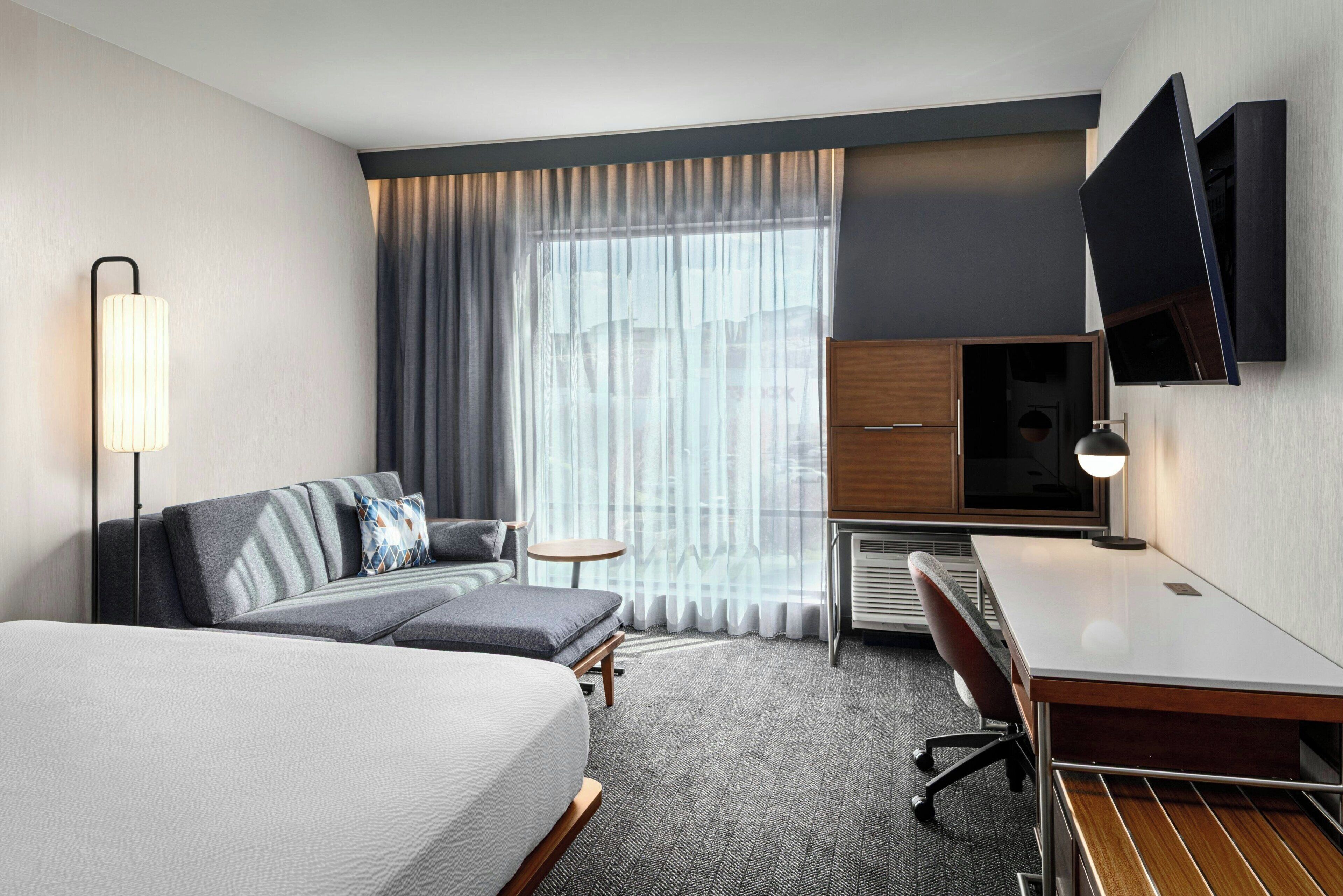 Quarto de hotel com cama, escrivaninha, cadeira, televisão e janela com cortinas.