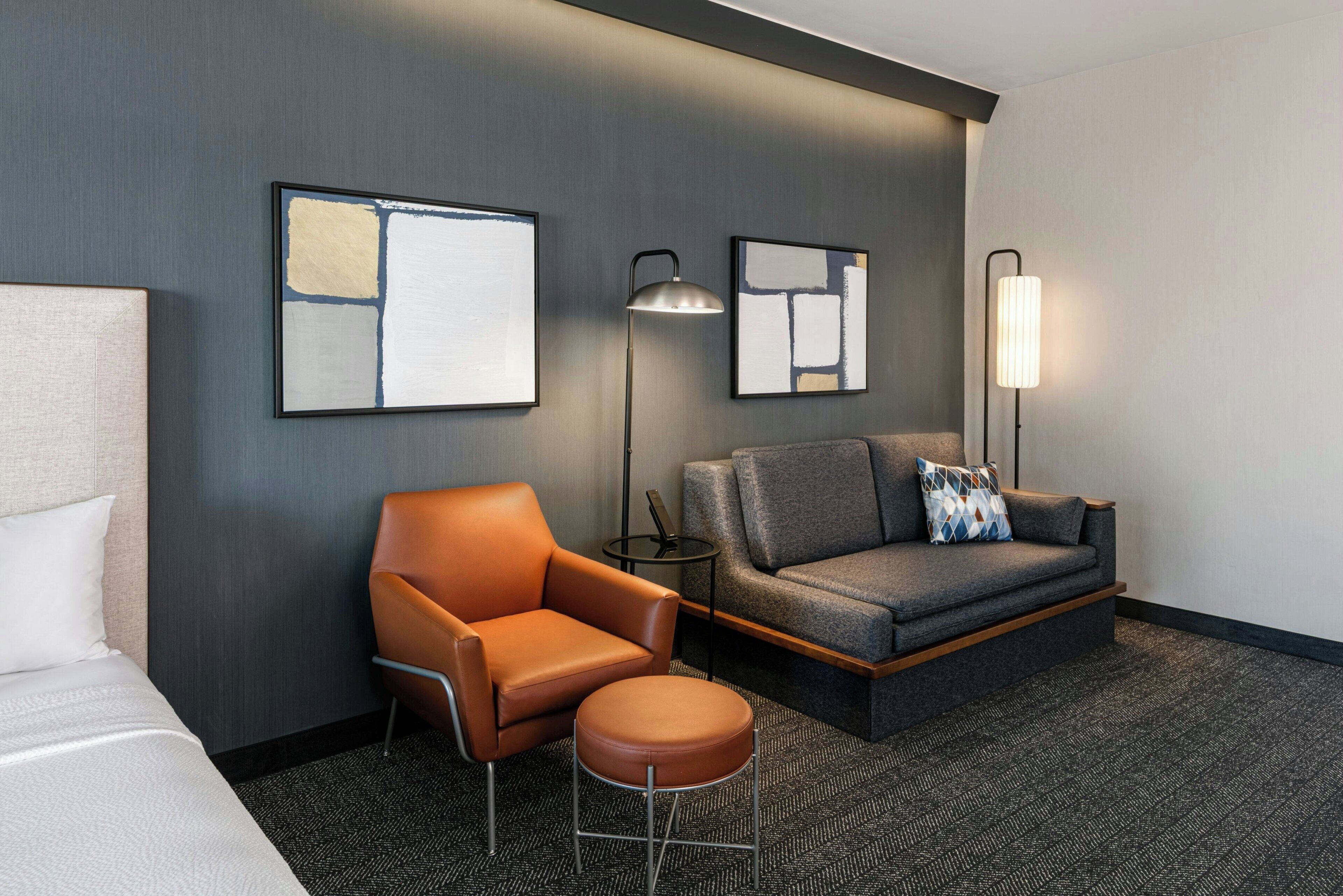 Una habitación de hotel con una cama, un sillón de cuero marrón, un sofá gris con un cojín estampado y dos lámparas fijadas a la pared.