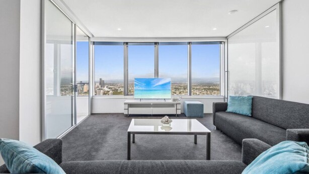 A modern living room with a large window, a flat-screen TV, a coffee table, and a gray sofa with blue cushions. - Sky Home on 54th Floor - Stunning South East Views (Surfers Paradise)
