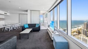 A modern living room with a large window, a sofa, a coffee table, and a TV. - Sky Home on 54th Floor - Stunning South East Views (Surfers Paradise)