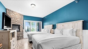 A bedroom with two beds, a stone fireplace, a TV, and a large window with a view of trees. - Breathtaking Lake Views | Studio, 2 Queen | Pools (Two Harbors)