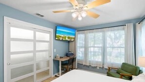 A bedroom with a bed, a desk, a chair, a ceiling fan, and a TV on the wall.