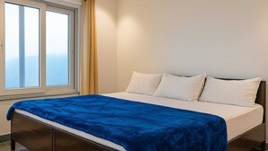 Un dormitori amb un llit, almoines blanques, un llençol blau i una finestra amb cortines.