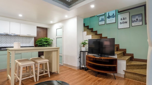 A modern kitchen with a herringbone backsplash, a dining area with stools, a TV on a wooden stand, and a staircase with framed pictures.