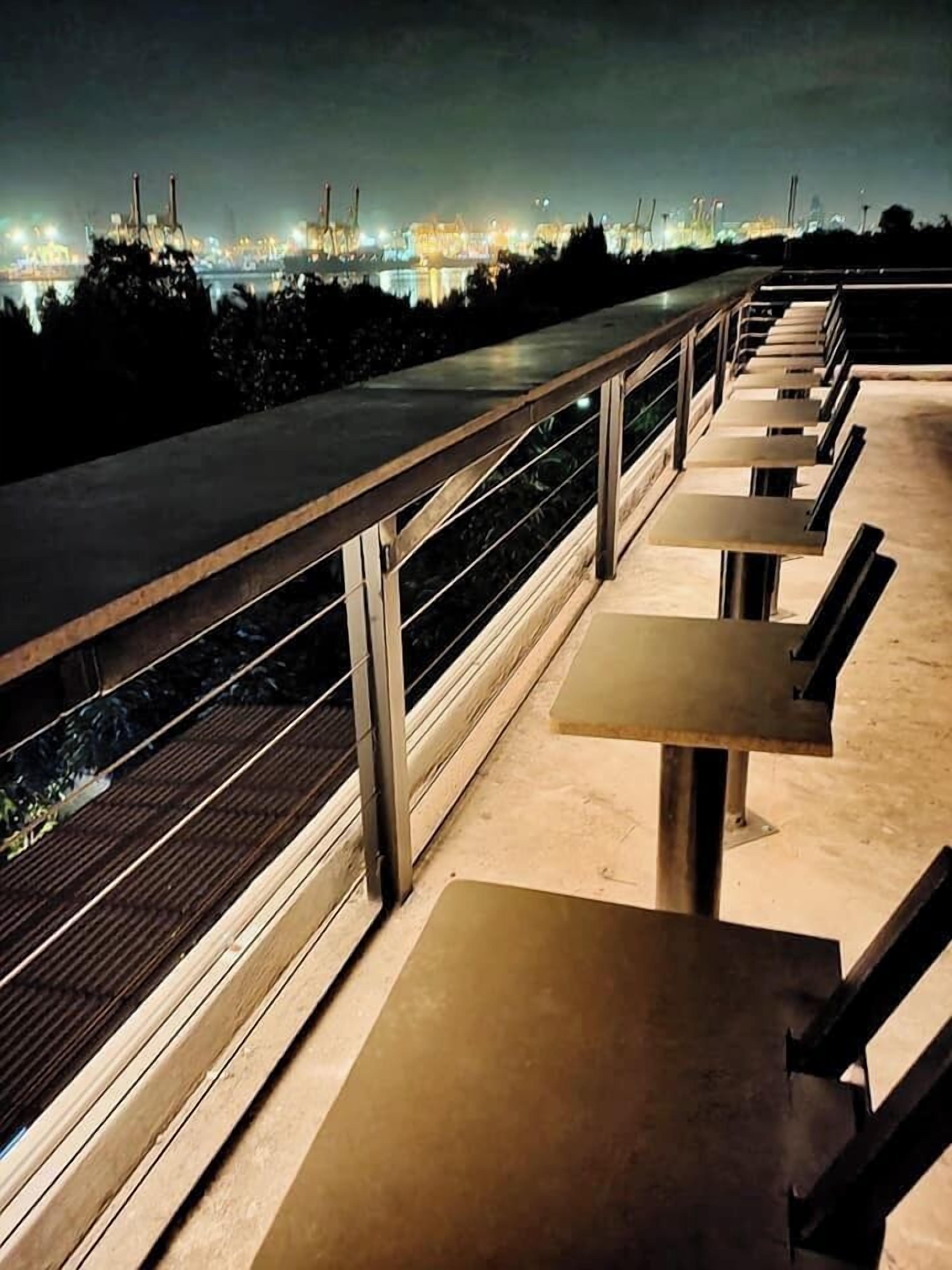 An outdoor seating area with metal railings and tables, overlooking a body of water and industrial structures in the background.