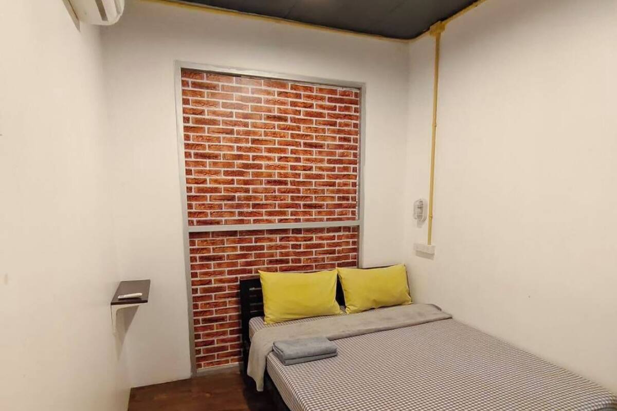 Habitació petita amb un llit individual, capçaleri de relleu de maó, sòl de fusta i paret blanca.
