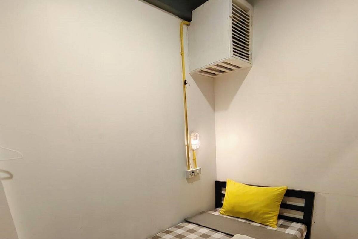 Habitació petita amb un llit individual, un edredó amb quadres, un coixí groc i aire condicionat fixat a la paret.