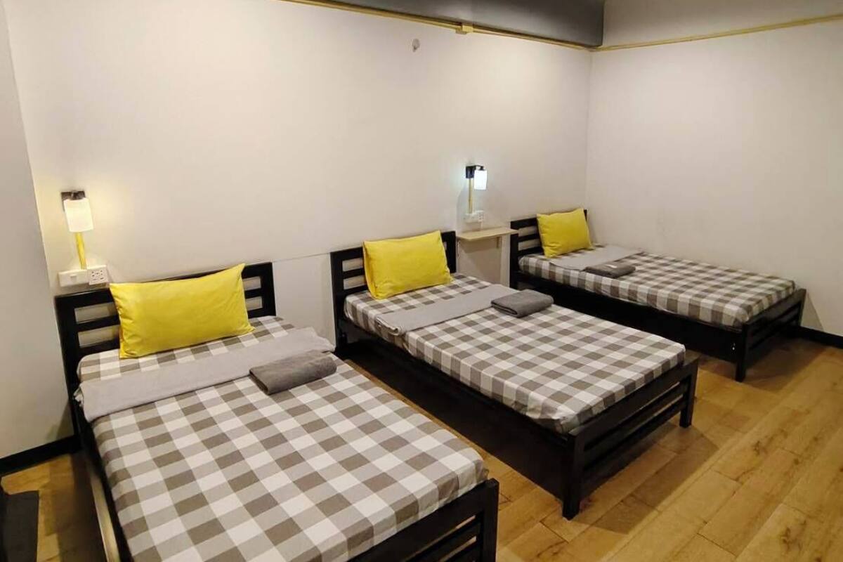 Habitació amb tres llits, cadascun amb un llençol escacat i una almoçada. A cada llit hi ha una nitòria amb una làmpada. La habitació té un paviment de fusta i un sostre blanc amb bigues al descobert.