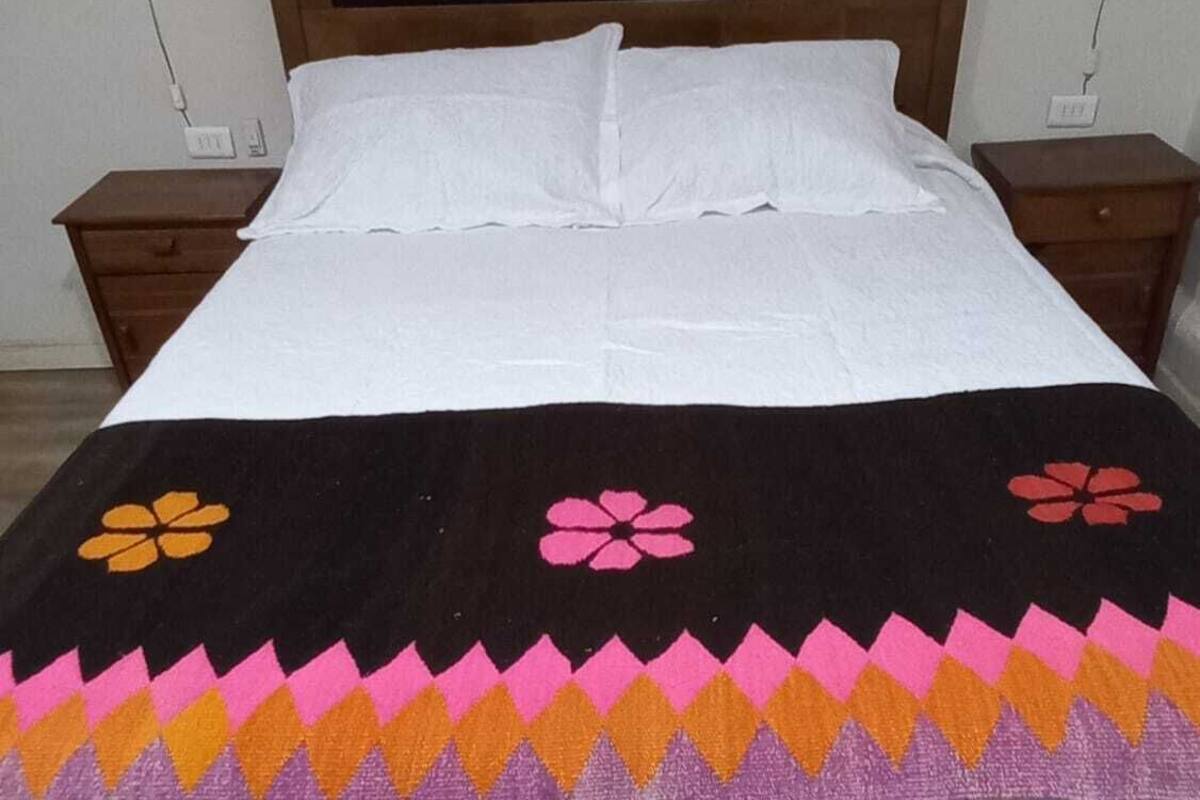 Een bed met een wit laken, een zwart-roze gebed en een kleurrijke wandtapijt erboven.