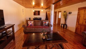 Un salon avec un plafond en bois, un canapé marron agrémenté de coussins colorés, une table basse ornée d’objets décoratifs et un téléviseur posé sur un meuble en bois.