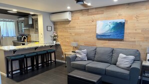 A modern living space with a kitchen area, grey sofa, and a wooden wall with a wave painting. - Dazzling Waikiki Apt Sleeps 7-excellent A/C & PARKING included (Honolulu)