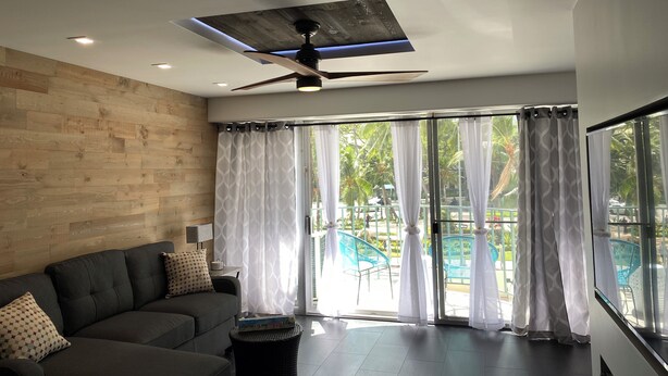 A modern living room with a grey sofa, a ceiling fan, and large windows with sheer curtains. - Dazzling Waikiki Apt Sleeps 7-excellent A/C & PARKING included (Honolulu)
