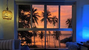 Une pièce dotée d’une grande fenêtre donnant sur une plage au coucher du soleil, d’un canapé, d’une petite table et d’un luminaire suspendu.