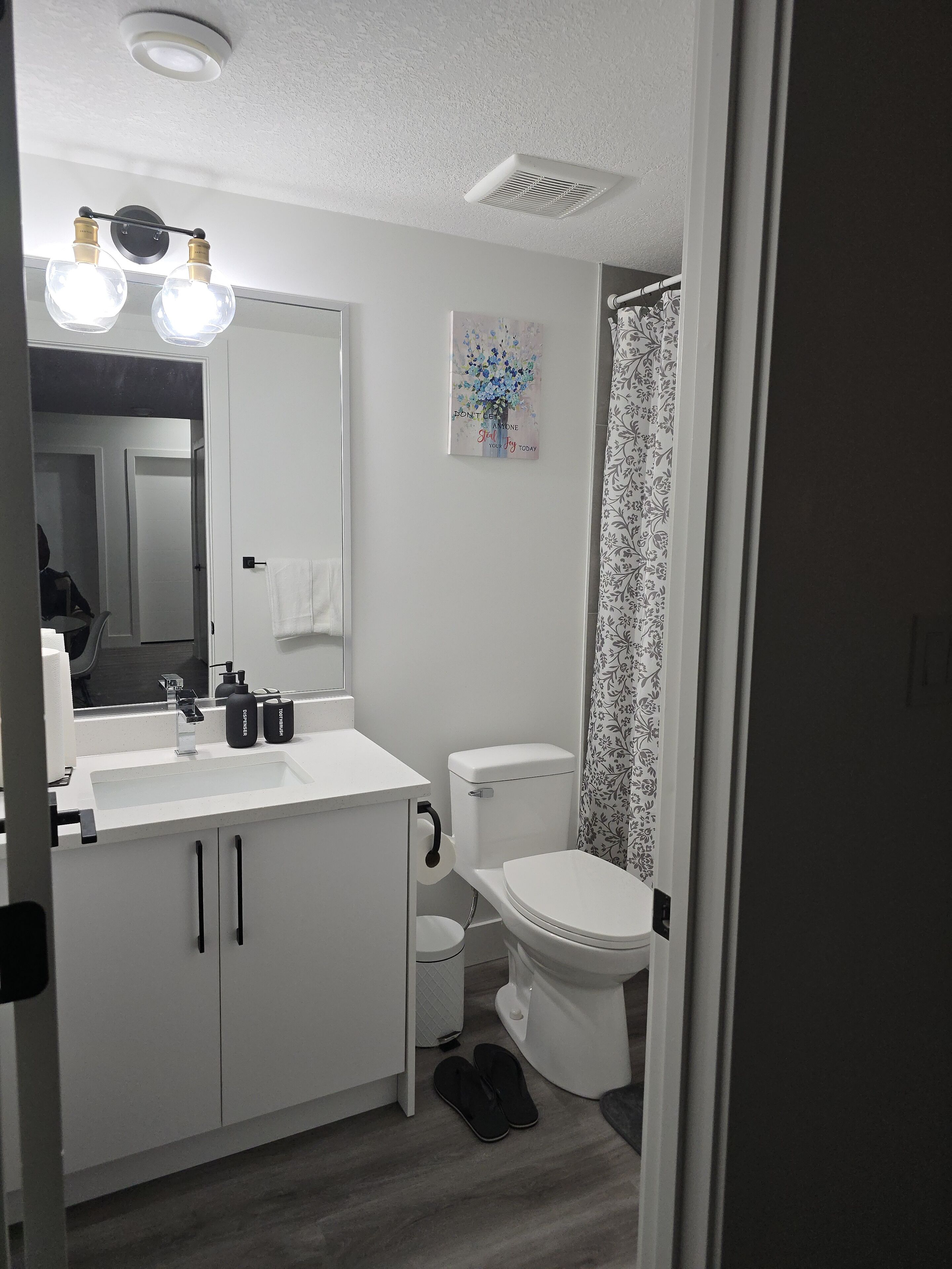 A modern bathroom with a white sink, mirror, and toilet. There is a shower curtain with a floral pattern, and a ceiling vent above the toilet.