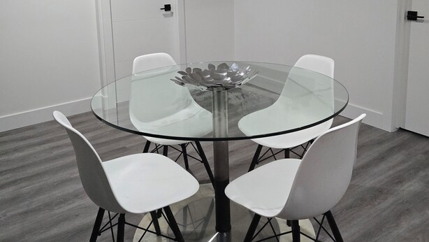 A round glass table with four white chairs, a white door, and a shelf with decorative items.