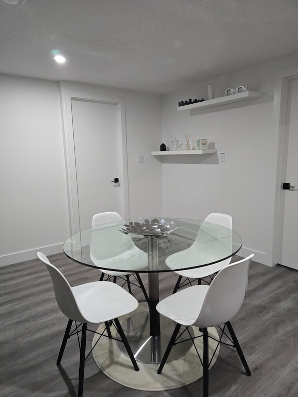A round glass table with four white chairs, a white door, and a shelf with decorative items.