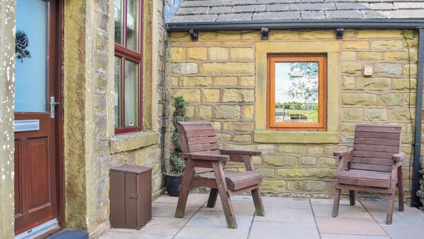 A stone cottage with a wooden door, two windows, and a small garden area with two wooden chairs.