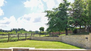 A stone wall with steps leading to a grassy area, a wooden fence, and trees in the background.