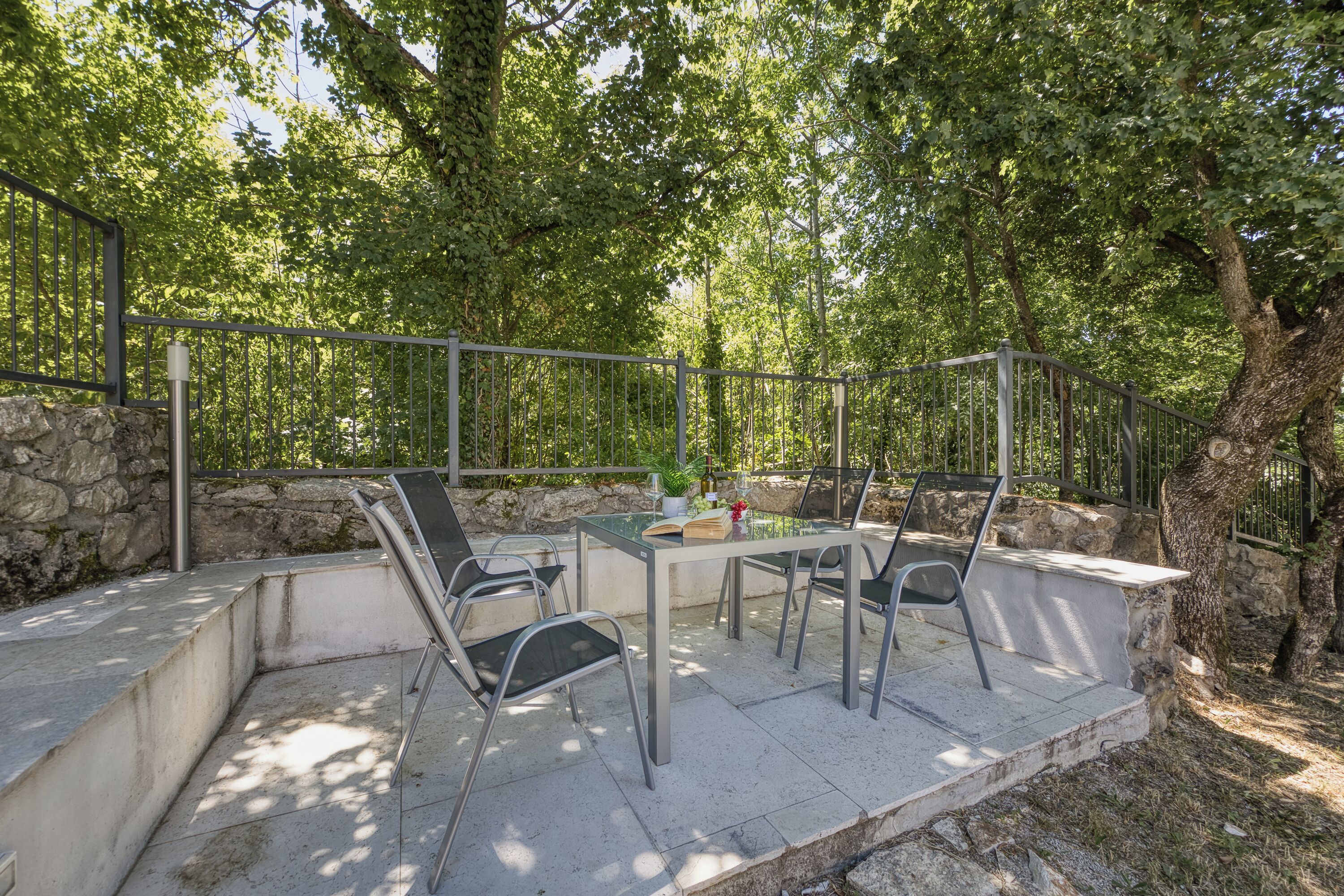 Um pátio externo com mesa e cadeiras, cercado por muros de pedra e árvores.