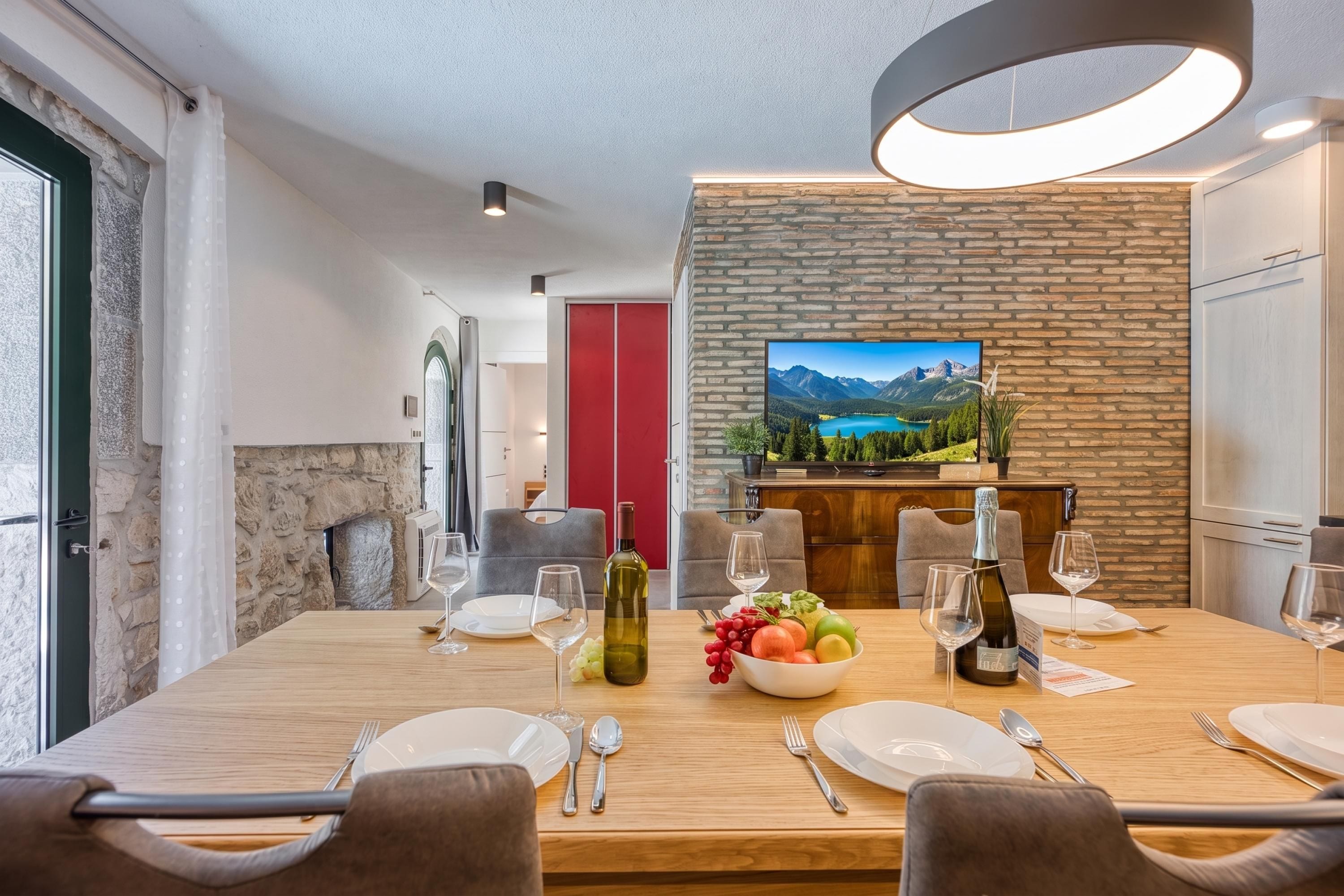 A dining area with a wooden table set for four, a television mounted on a brick wall, and a stone fireplace.