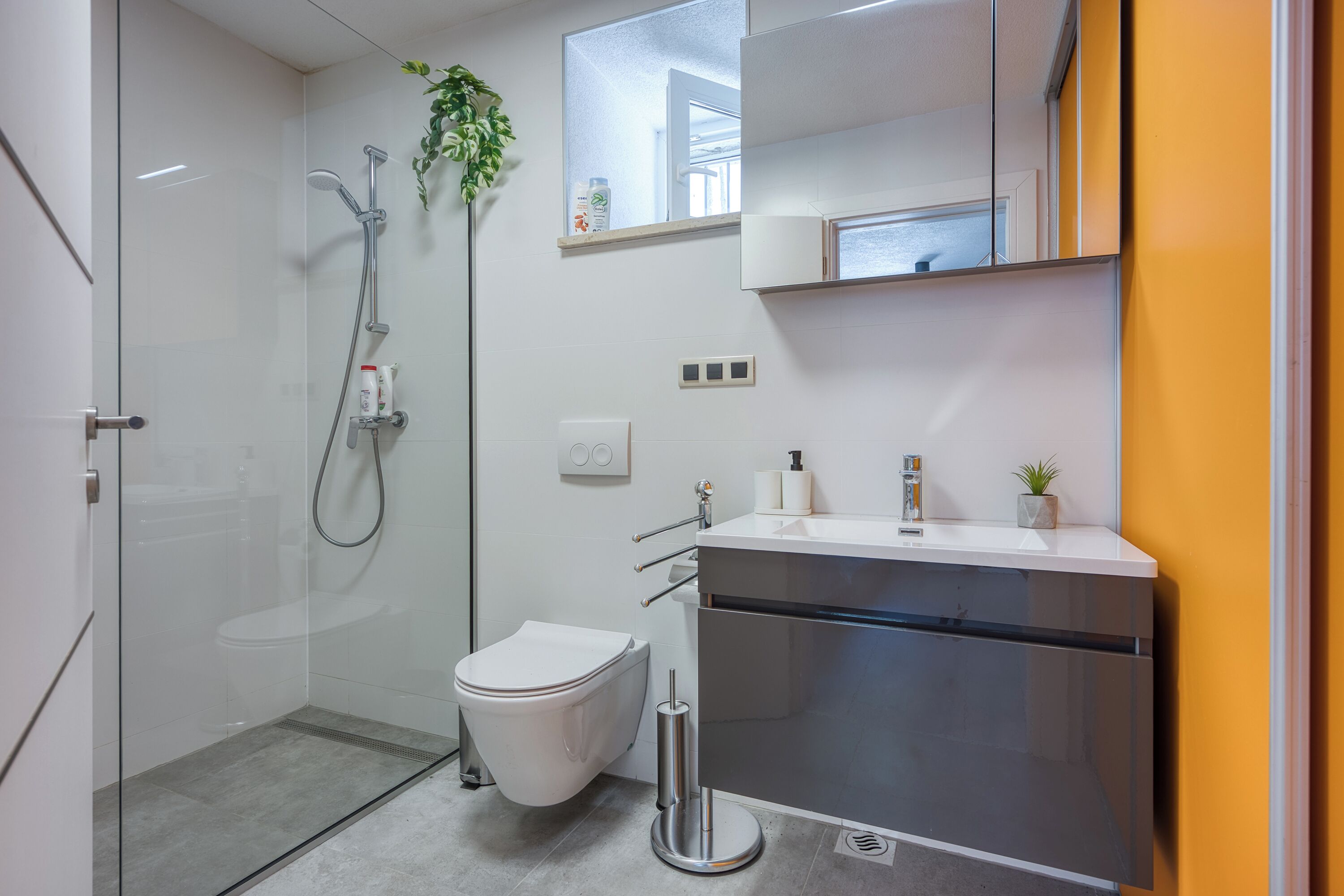 Un baño moderno con cabina de ducha de vidrio, inodoro blanco y lavamanos de acero inoxidable con espejo superior.