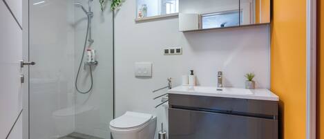 Un baño moderno con cabina de ducha de vidrio, inodoro blanco y lavamanos de acero inoxidable con espejo superior.