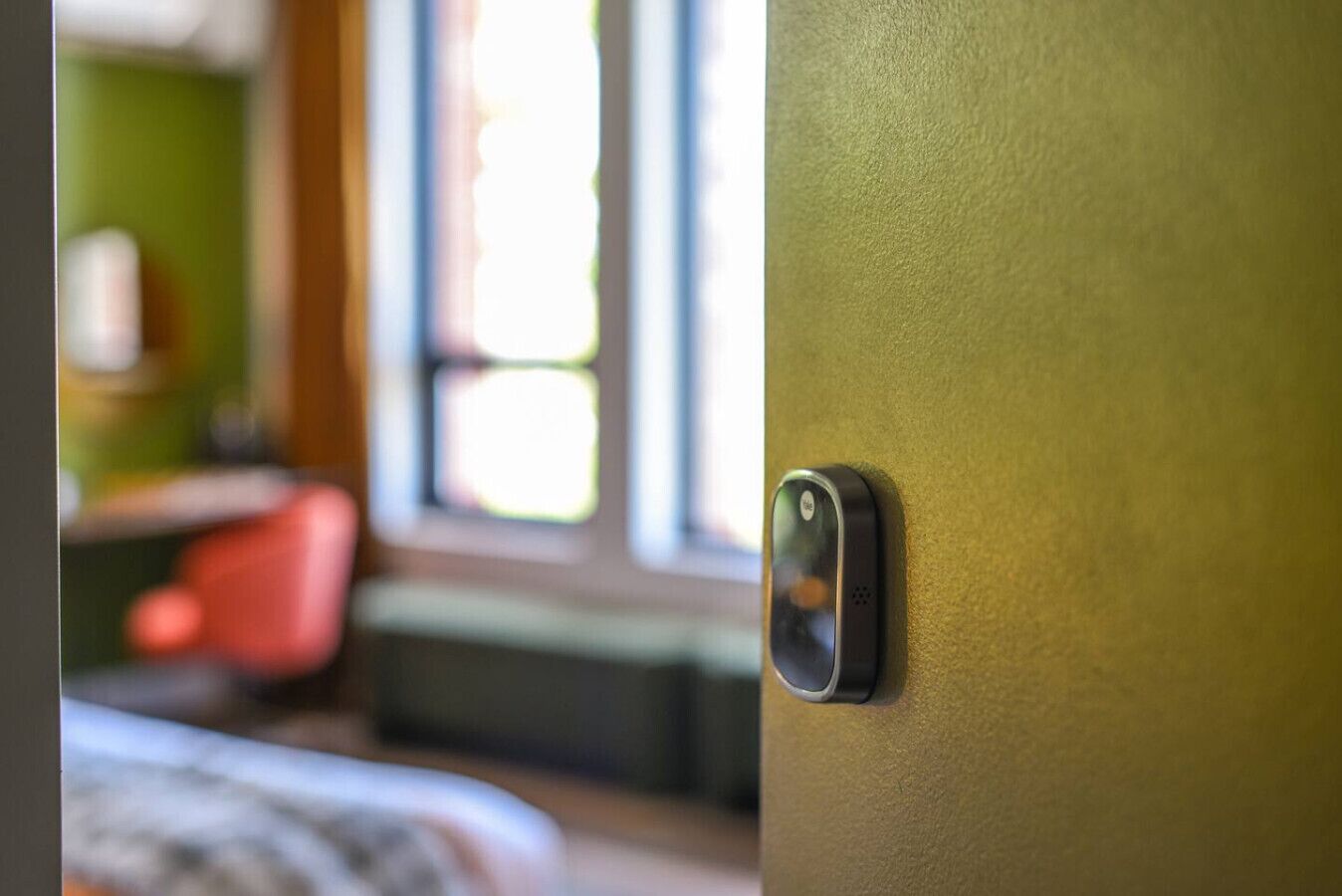 A door with a modern intercom system, a window with blinds, and a glimpse of a room with a bed and a red chair.