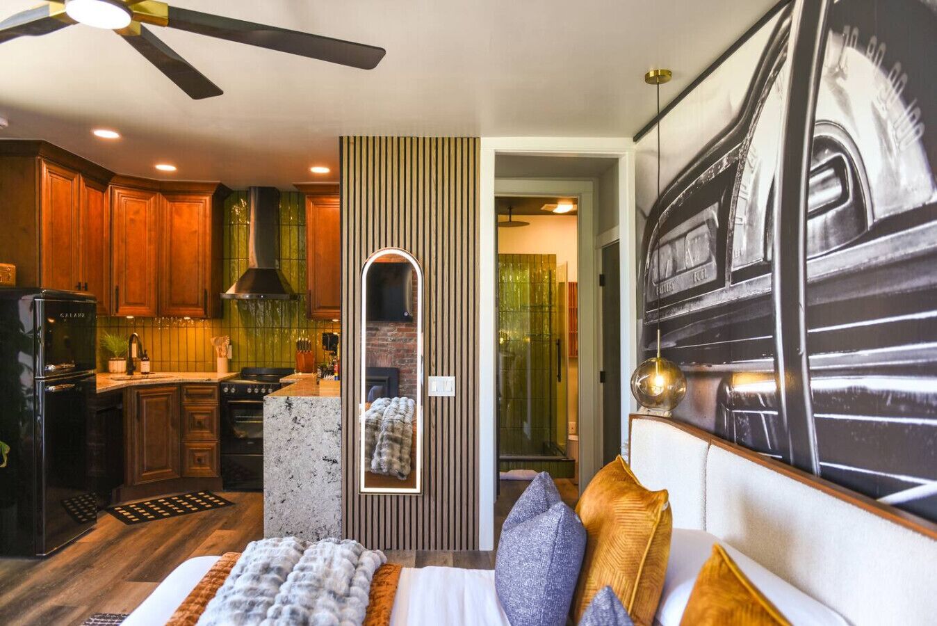 A modern hotel room with a large bed, a sitting area with a sofa and pillows, a kitchen with wooden cabinets and a black refrigerator, and a dining area with a table and chairs.