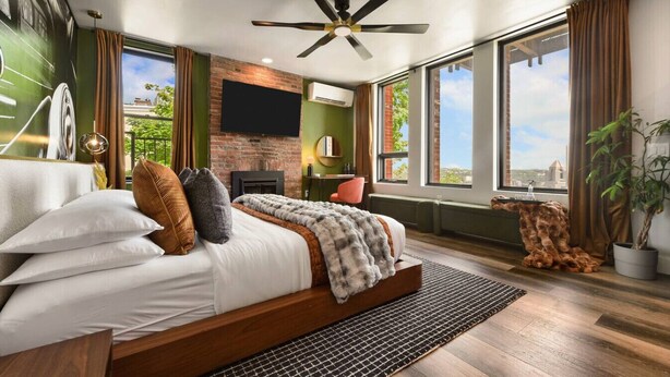 Une chambre moderne dotée d’un grand lit, d’un ventilateur de plafond, d’une cheminée et de grandes fenêtres offrant une vue sur la ville.