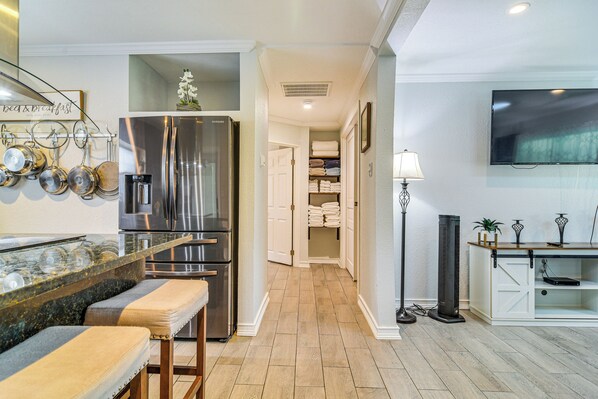 A modern kitchen with a stainless steel refrigerator, a marble countertop, and a bench. There is a hallway leading to a closet with folded towels.