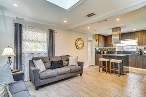 A modern living room with a grey sofa, a coffee table, and a lamp. There is a kitchen area with a bar and stools.