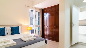 A hotel room with a bed, a window with curtains, a wooden wardrobe, and a door leading to another room.