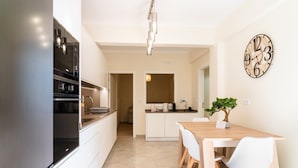 A modern kitchen with white cabinets, a stainless steel refrigerator, and a dining area with a wooden table and white chairs. A clock is mounted on the wall, and a small potted plant is on the table.