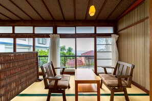 A traditional Japanese room with tatami flooring, a low wooden table, two wooden chairs, and a view through large windows.