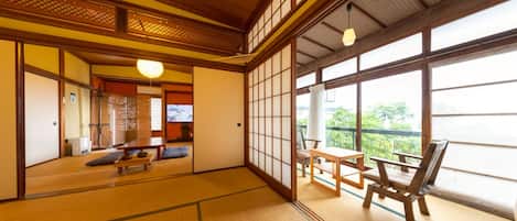 A traditional Japanese room with tatami flooring, a sliding door, a view of greenery outside, and a table with chairs.