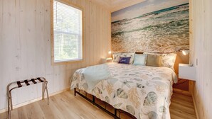 A bedroom with a wooden ceiling, a large ocean mural, a bed with a seashell-themed bedspread, a bedside table, and a window with blinds.