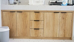 Een keuken met een witte tegelachtergrond, een houten kastje met twee lades, een witte spoelbak en een koffiezetapparaat op het aanrecht.