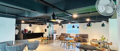 Una cafetería moderna con diversas opciones de asientos, incluyendo sillas y mesas, un ventilador grande y una zona de mostrador.