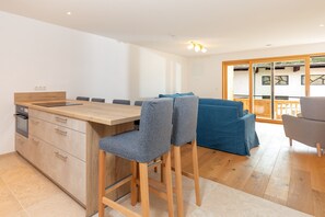 Une cuisine moderne avec un plan de travail en bois et des tabourets de bar, un coin repas avec un canapé bleu et, vue à travers une porte vitrée, une terrasse extérieure.