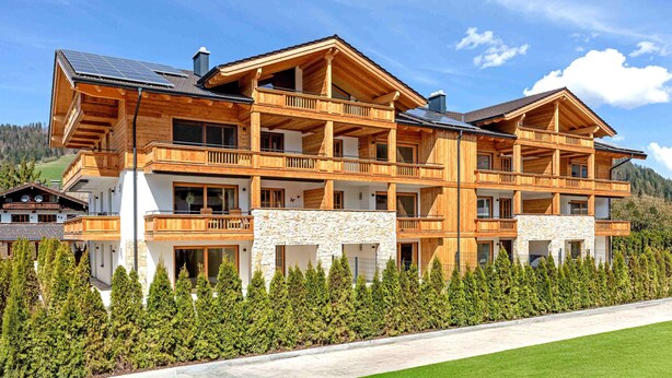 Un chalet en bois à plusieurs étages, doté de balcons, d’éléments décoratifs en pierre et offrant une vue sur la forêt environnante.